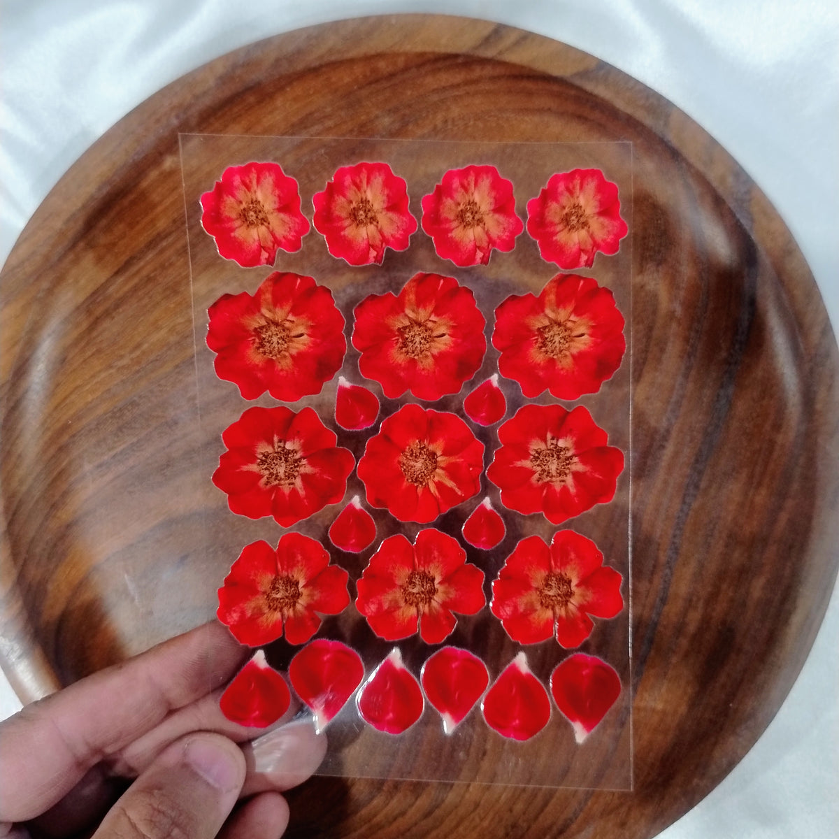 A sheet of 3D UV DTF transfer stickers featuring realistic red rose flowers and individual rose petals held over a wooden plate for resin art and crafts.