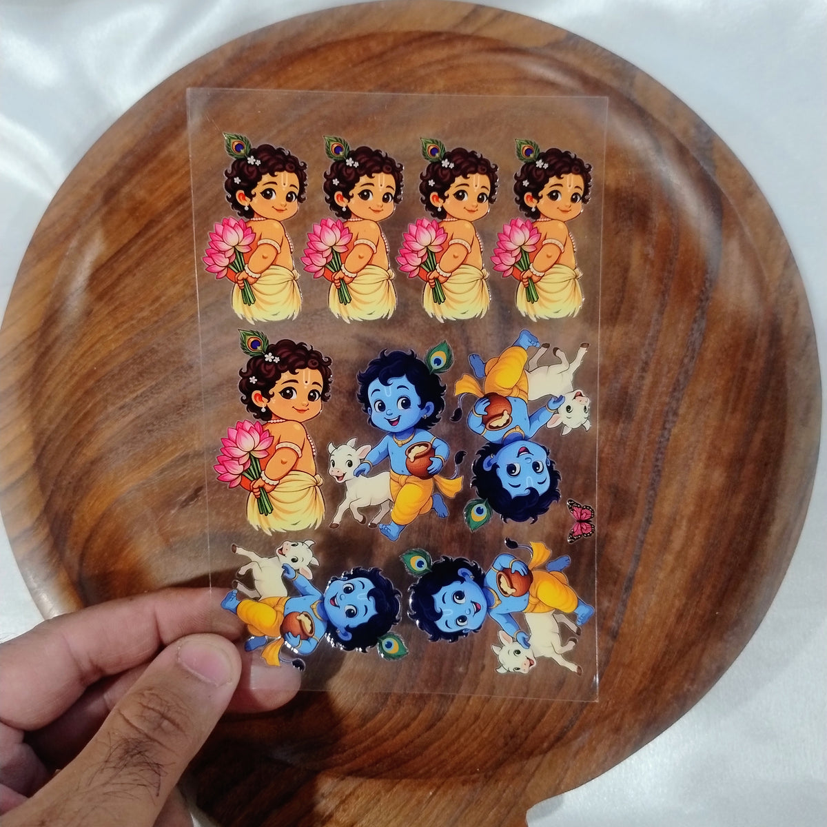 A sheet of 3D cute cartoon Little Krishna UV DTF transfer stickers holding pink lotus bouquets and playing with calves held over a wooden plate.