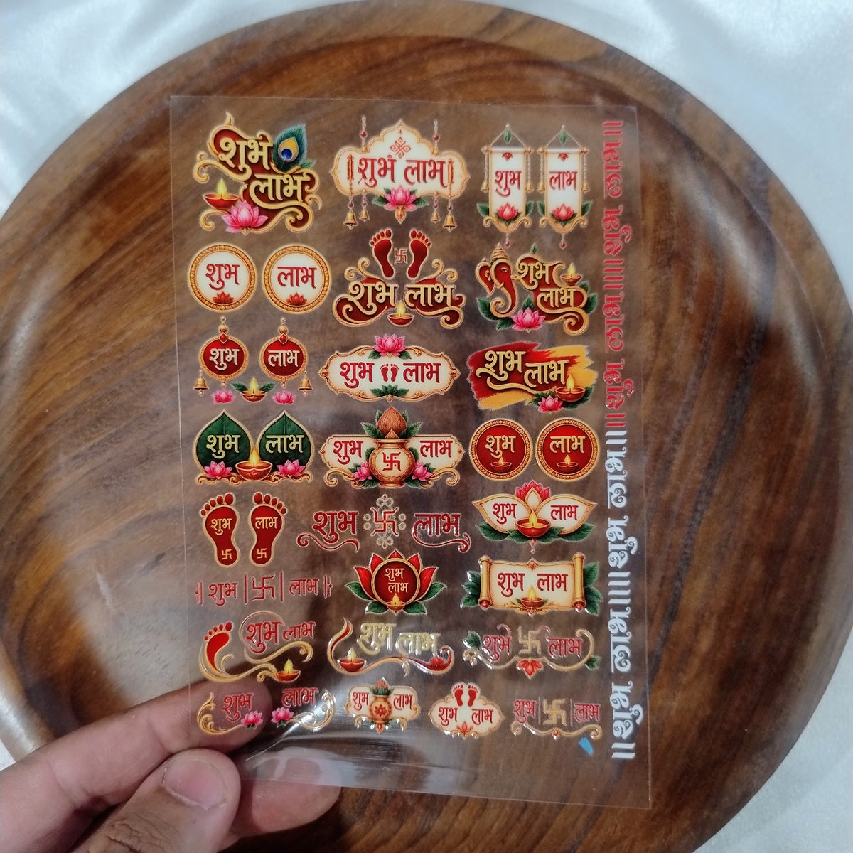 A sheet of 3D designer Shubh Labh UV DTF transfer stickers with peacock feather, kalash, diya, and lotus motifs held over a wooden plate.