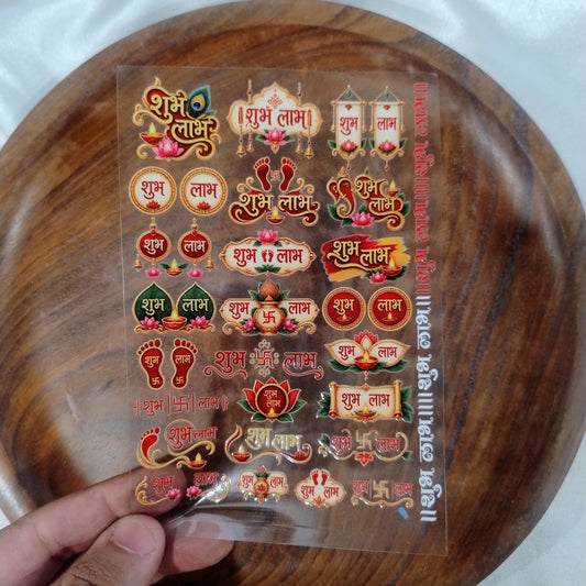 A sheet of 3D designer Shubh Labh UV DTF transfer stickers with peacock feather, kalash, diya, and lotus motifs held over a wooden plate.