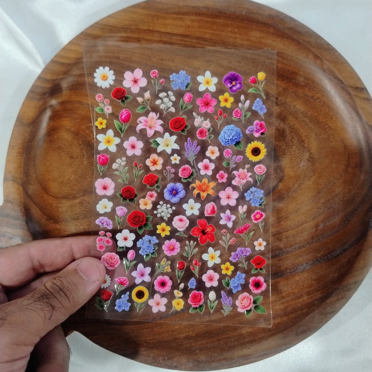 A sheet of colorful mini 3D floral UV DTF transfer stickers with roses, sunflowers, and lavender designs held over a wooden bowl for resin art and candle jar crafts