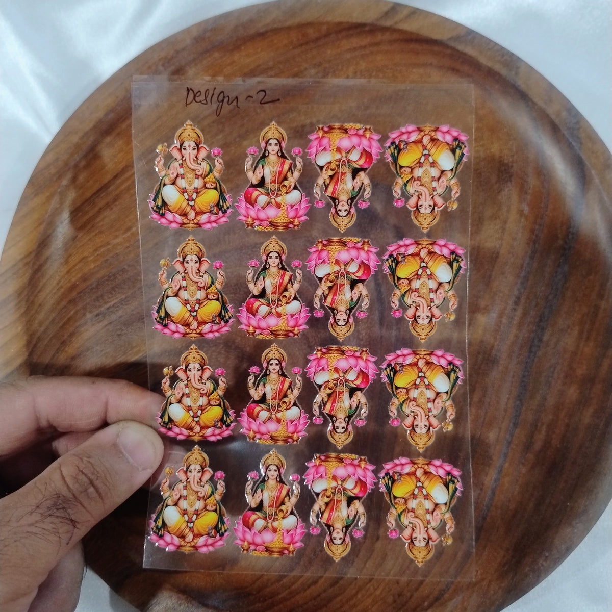 A sheet of 3D Lord Ganesha and Goddess Lakshmi UV DTF transfer stickers seated on lotus flowers held over a wooden plate for resin art and candle jar crafts