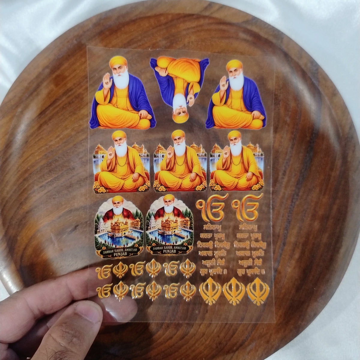 A sheet of 3D Guru Nanak Dev Ji, Golden Temple Amritsar, Ik Onkar, and Khanda UV DTF transfer stickers held over a wooden plate for resin art and crafts.