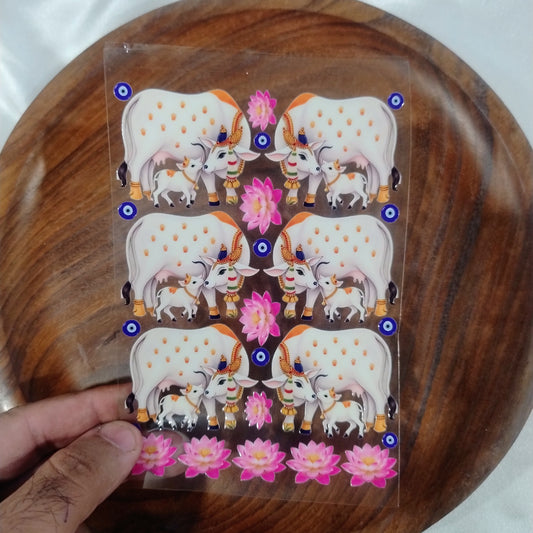 A sheet of 3D Kamdhenu cow and calf UV DTF transfer stickers with lotus flowers and blue evil eye symbols held over a wooden plate for resin art and crafts.