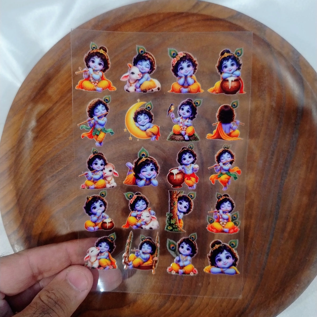 A sheet of colorful cute Little Krishna UV DTF transfer stickers with Bal Gopal playing flute and eating butter, held over a wooden plate for resin art and candle jar crafts.