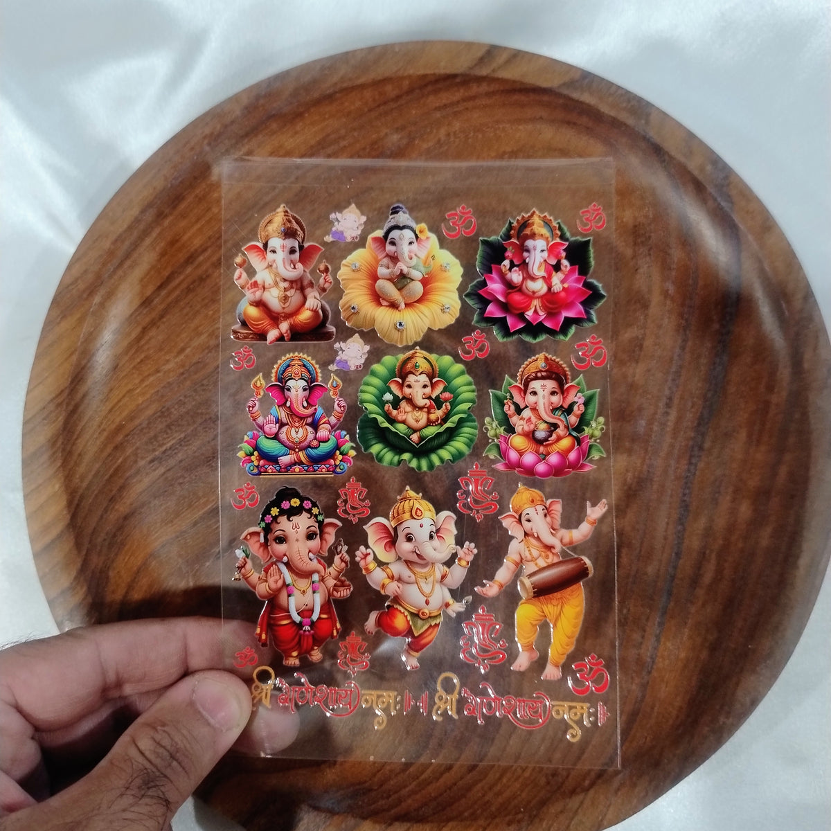 A sheet of 3D colorful Lord Ganesha UV DTF transfer stickers on lotus flowers with Shree Ganeshay Namah text held over a wooden plate.