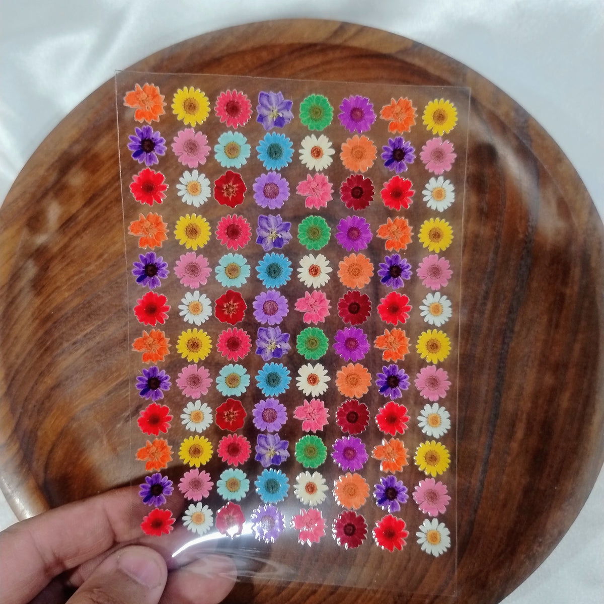 A sheet of colorful mini daisy and sunflower UV DTF transfer stickers with a pressed flower effect held over a wooden plate for resin art and crafts
