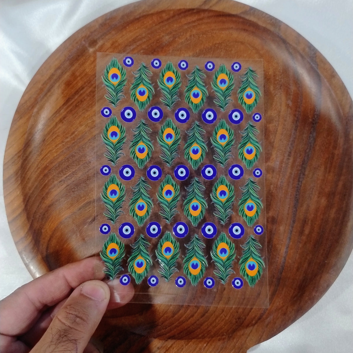 A sheet of 3D glossy peacock feather and blue evil eye UV DTF transfer stickers held over a wooden plate for resin art and candle jar crafts.