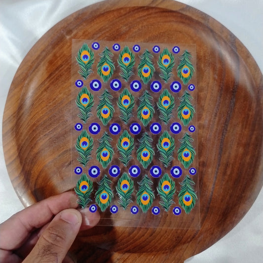 A sheet of 3D glossy peacock feather and blue evil eye UV DTF transfer stickers held over a wooden plate for resin art and candle jar crafts.