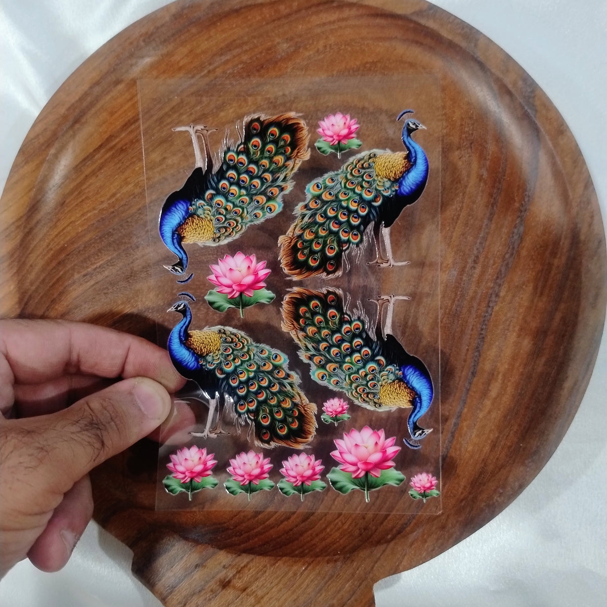 A sheet of 3D vibrant blue peacock UV DTF transfer stickers with blooming pink lotus flowers held over a wooden plate for resin art and candle jar crafts.