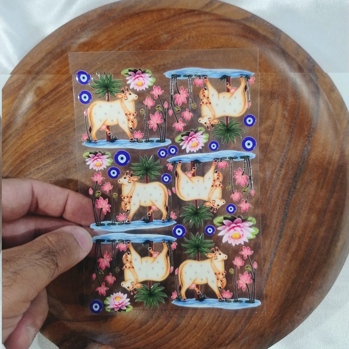 A sheet of 3D Pichwai style holy cow UV DTF transfer stickers standing in a lotus pond with palm trees and blue evil eye symbols held over a wooden plate.