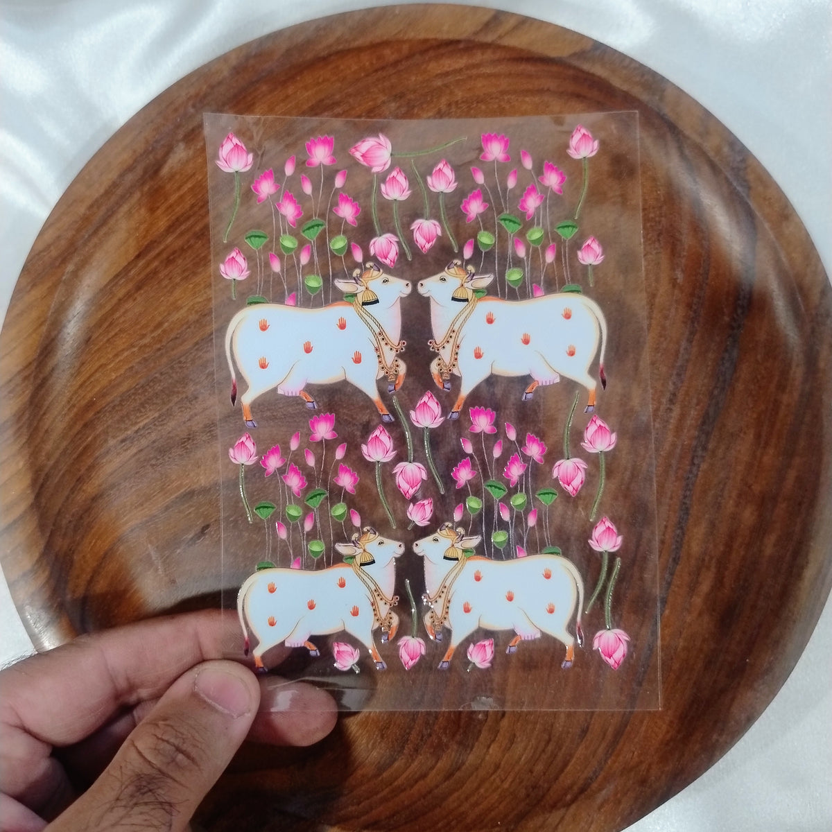 A sheet of 3D traditional Pichwai sacred cow UV DTF transfer stickers surrounded by blooming pink lotus stems held over a wooden plate.