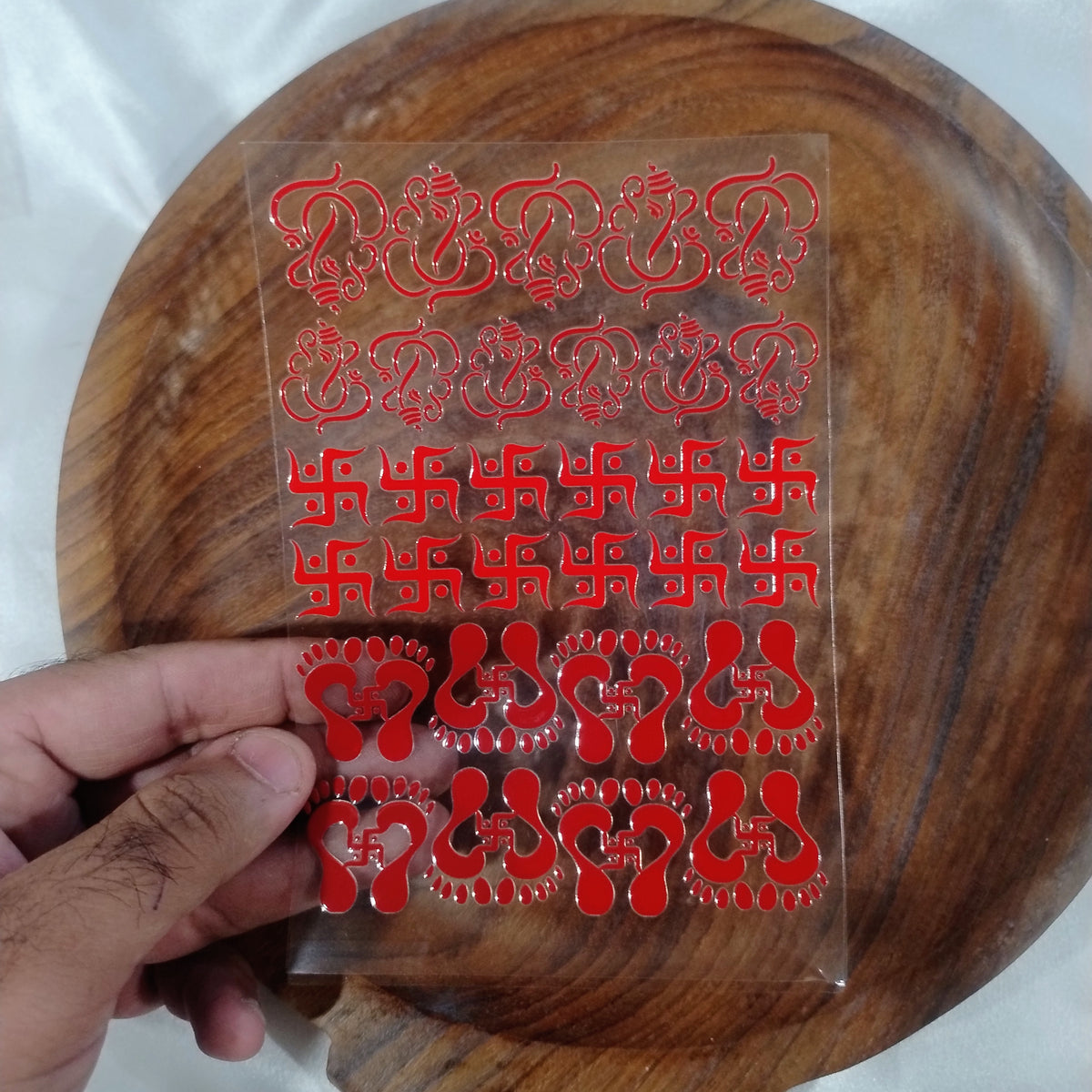 A sheet of 3D red glossy UV DTF transfer stickers featuring Lord Ganesha, Swastik symbols, and Laxmi Charan footprints held over a wooden plate for resin art and crafts.