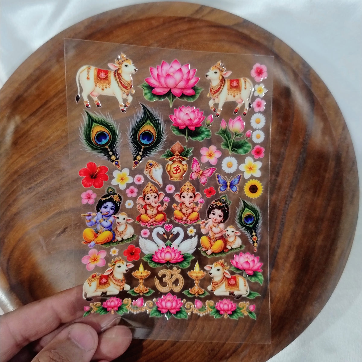 A sheet of 3D colorful UV DTF transfer stickers featuring Little Krishna, Lord Ganesha, Kamdhenu cows, peacock feathers, and lotus flowers held over a wooden plate