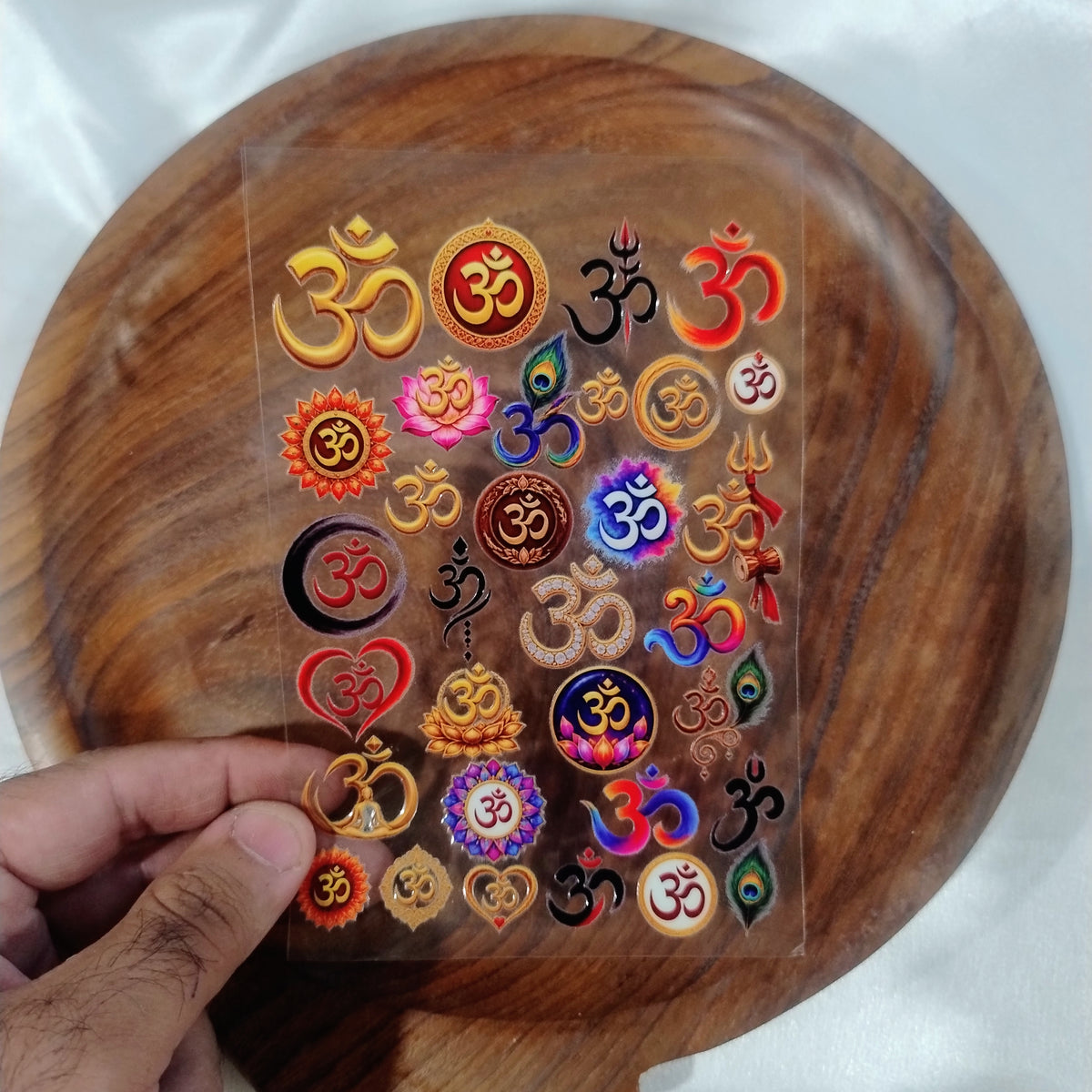 A sheet of 3D colorful and metallic gold Om symbol UV DTF transfer stickers with lotus and peacock feather accents held over a wooden plate