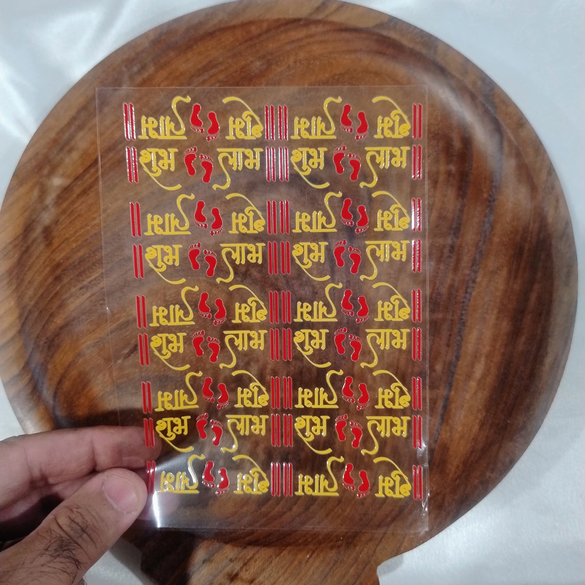 A sheet of 3D red and gold Shubh Labh calligraphy UV DTF transfer stickers with Laxmi Charan footprints held over a wooden plate for resin art and door decor