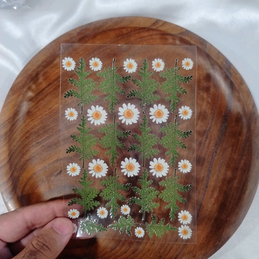 A sheet of 3D UV DTF transfer stickers featuring realistic white daisies and green fern leaves held over a wooden plate for resin art and crafts
